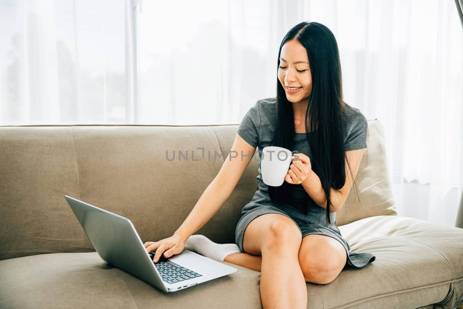 Woman on sofa sips coffee watches laptop education in cozy living room by Sorapop