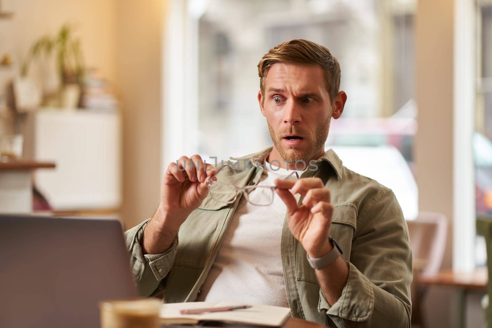 Portrait of man in cafe, takes off his glasses, looks shocked and concerned at his laptop screen, reading upsetting news.