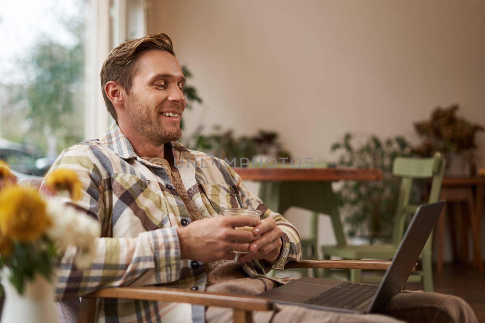 Image of handsome businessman working from cafe, drinking coffee and doing online project on his laptop, smiling and looking happy, finished his freelance task, looking pleased.