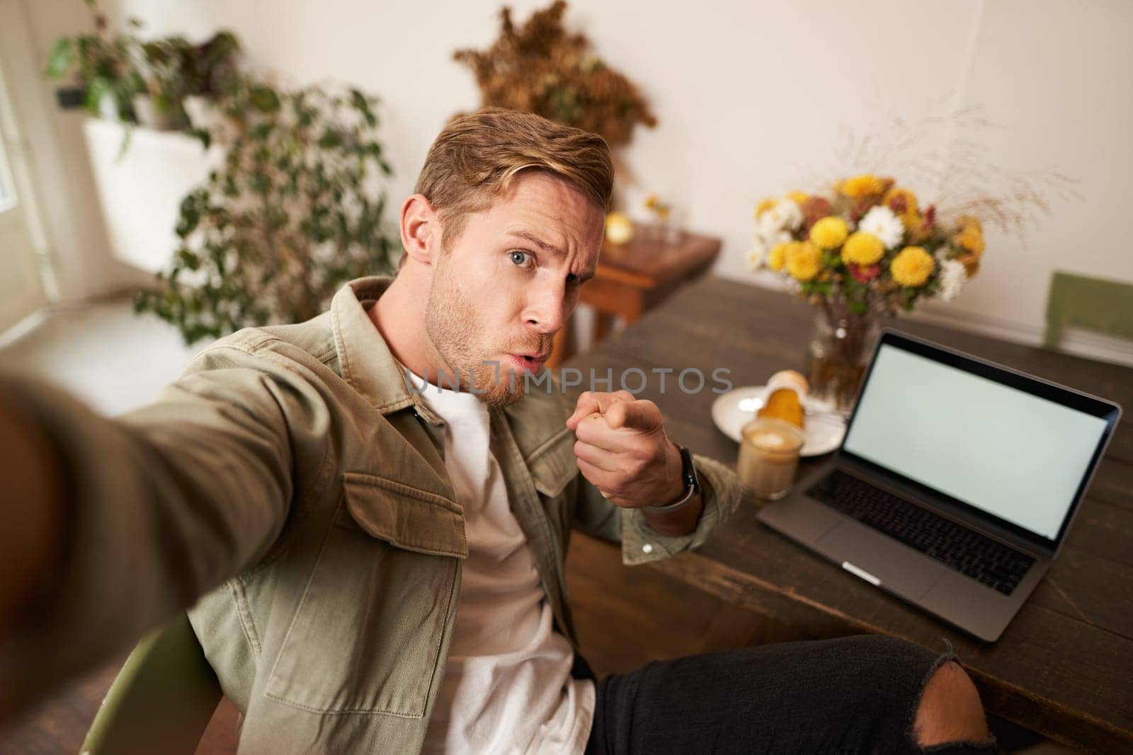 Portrait of serious man takes selfie with fist and threatening face, sits in cafe, poses in coffee shop in front of table with laptop and glass of coffee.