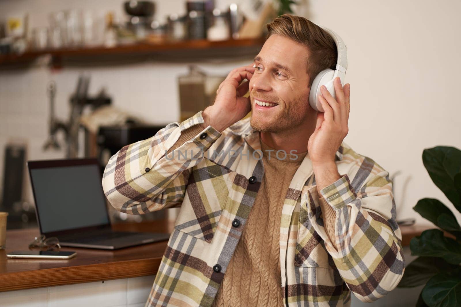 Portrait of handsome happy guy, freelancer working and listening to music in wireless headphones, dancing on his chair in cafe, enjoying favourite song in earphones.