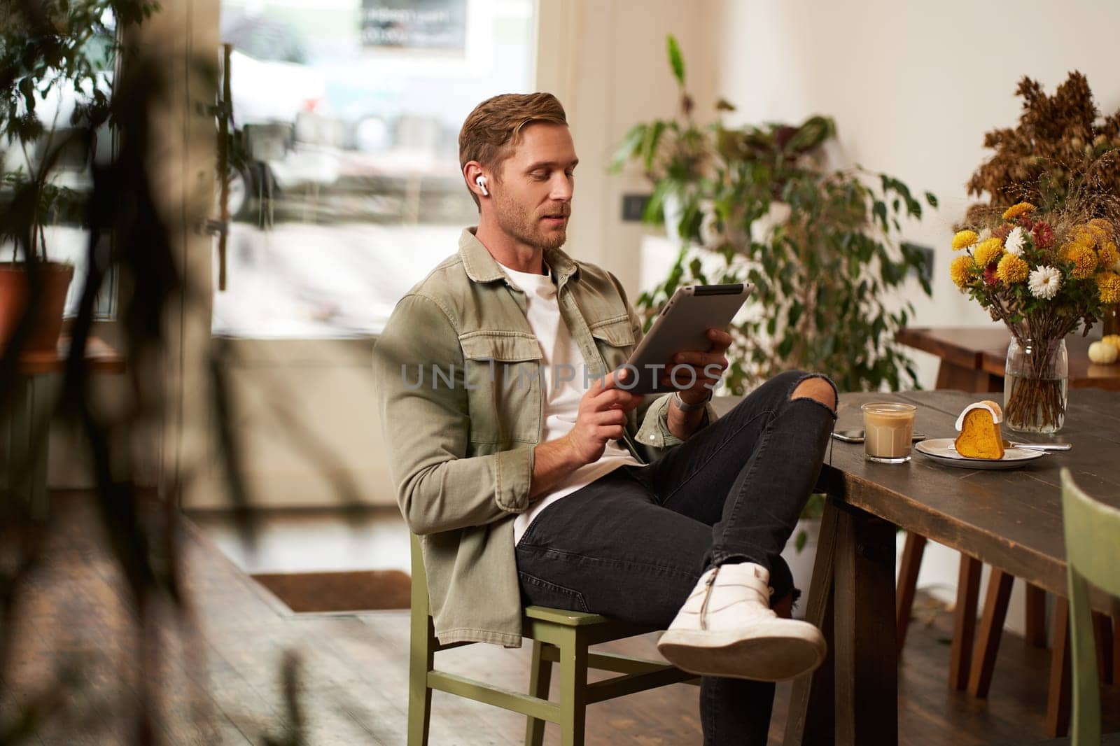 Lifestyle shot of handsome young man sitting in a cafe in front of the table, drinking coffee, wearing wireless headphones, listening to music or watching video in public space, using digital tablet.