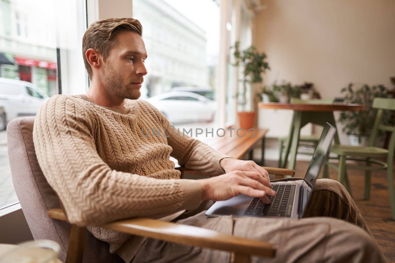 Working people and public space concept. Young handsome man, ux ui designer sitting with laptop in cafe, drinking cappuccino, looking at screen with serious face.