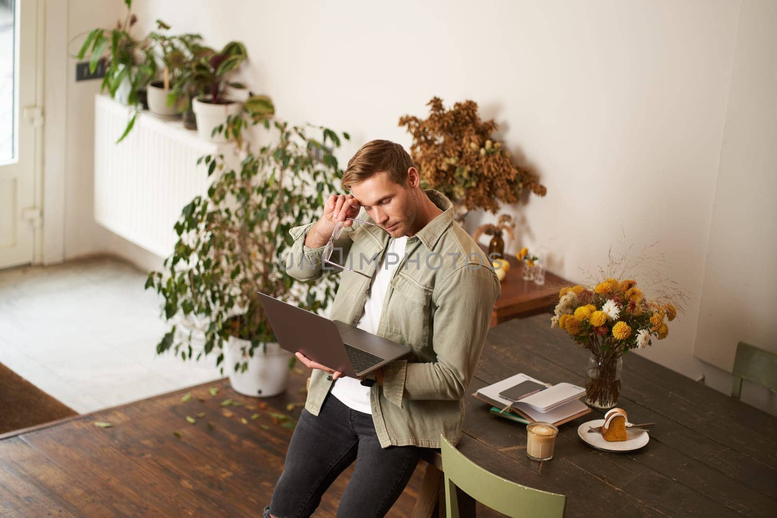 Image of young man, successful businessman sitting on table, looking at laptop, working in an office, looking concentrated on the project, doing job task, drinking coffee.