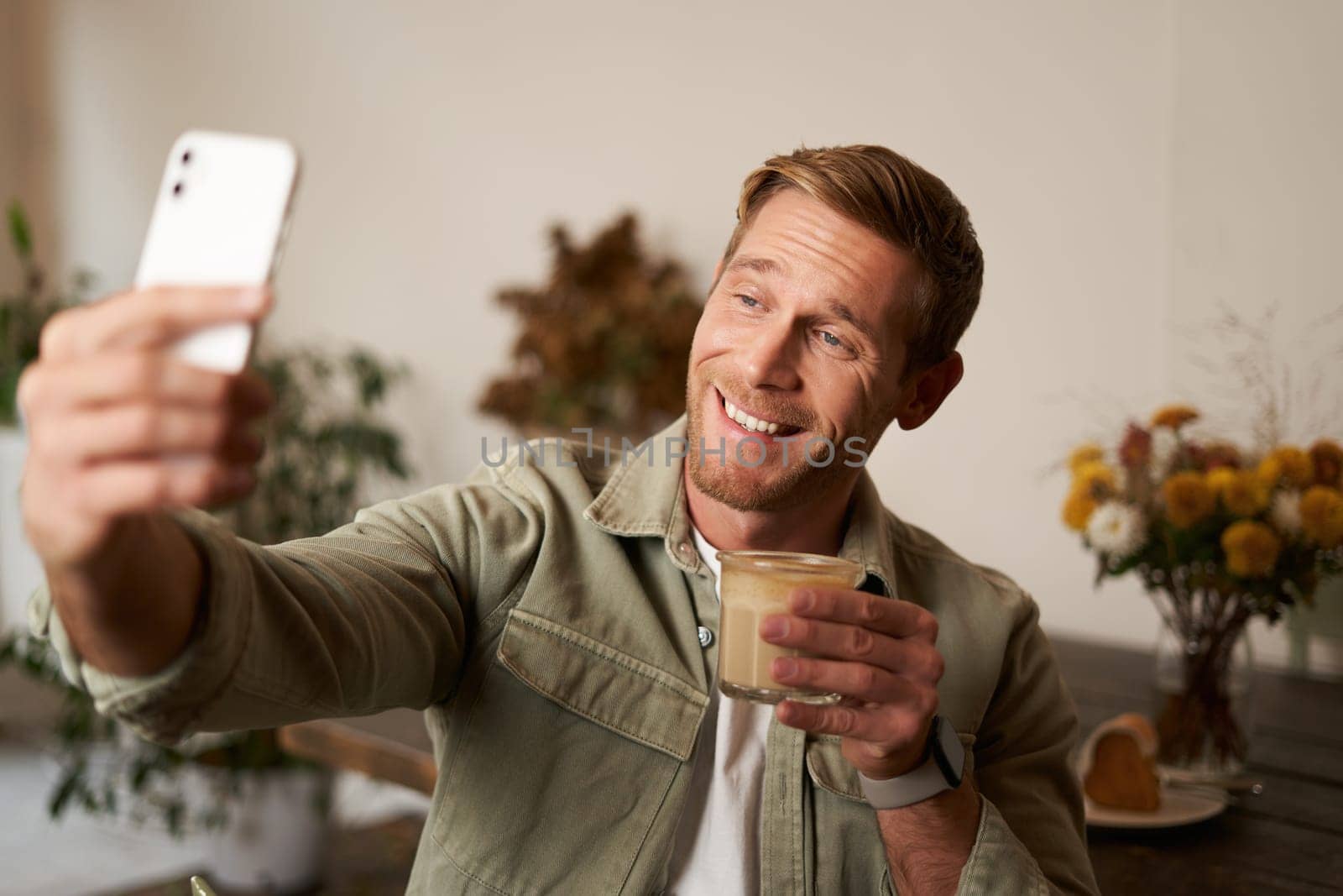 Portrait of smiling handsome young man, video chats with friend from cafe, doing selfies with cup of coffee.