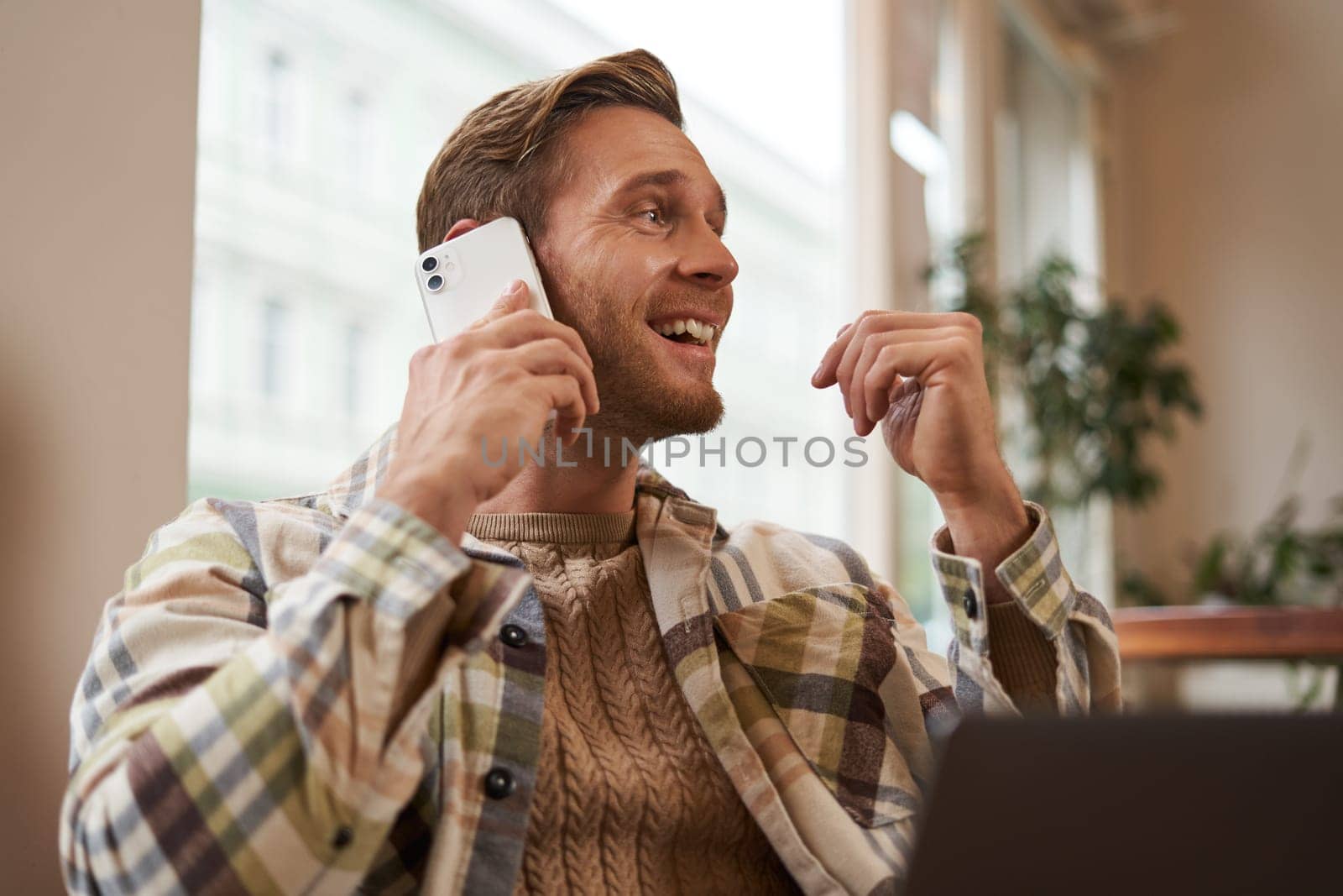 Portrait of young businessman, freelancer working remotely from coffee shop in city, sitting in chair with laptop, answer a phone call, talking on mobile to someone.