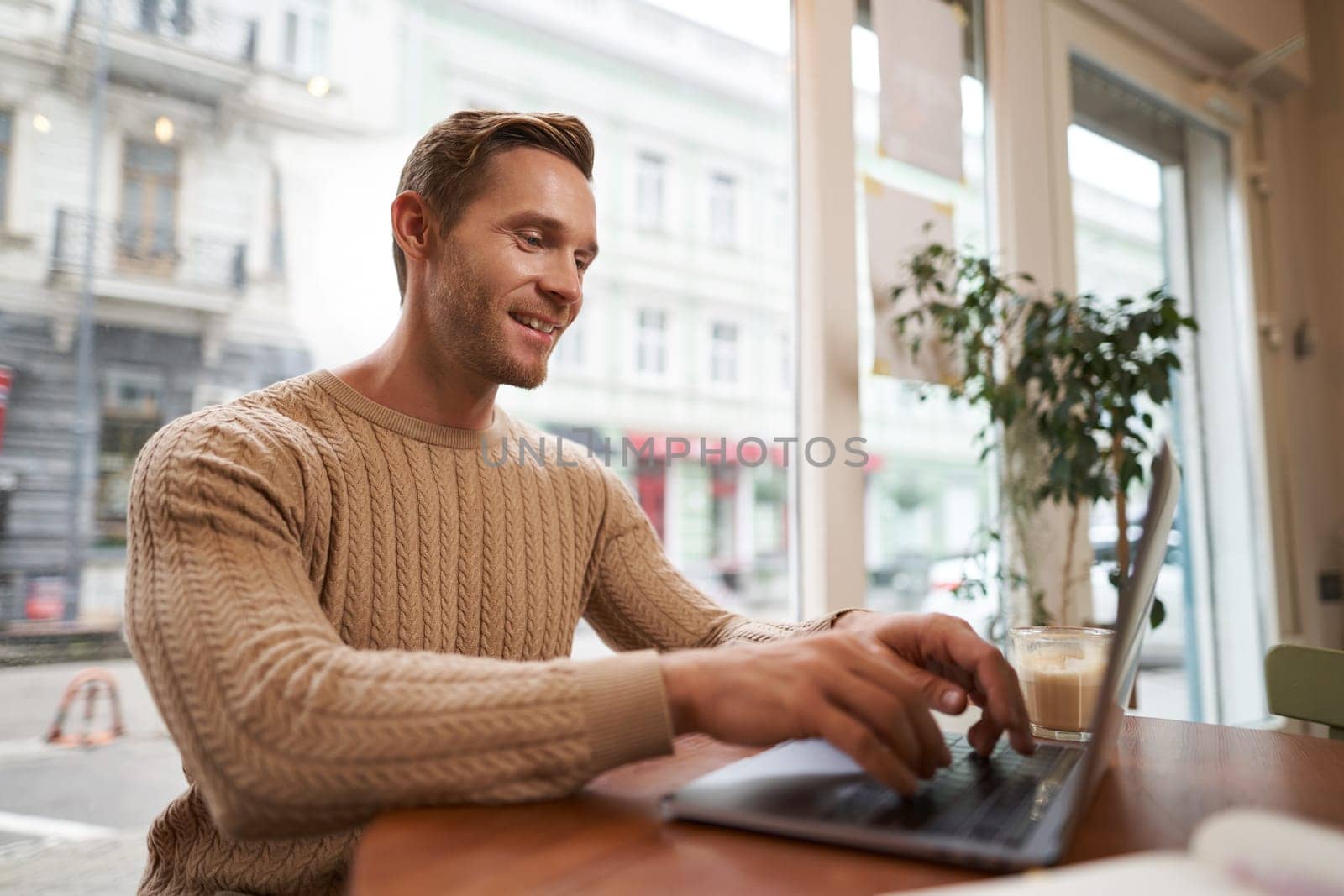 Portrait of handsome young man sitting in cafe with laptop. Digital nomad, freelancer working outdoors from coffee shop, typing something.