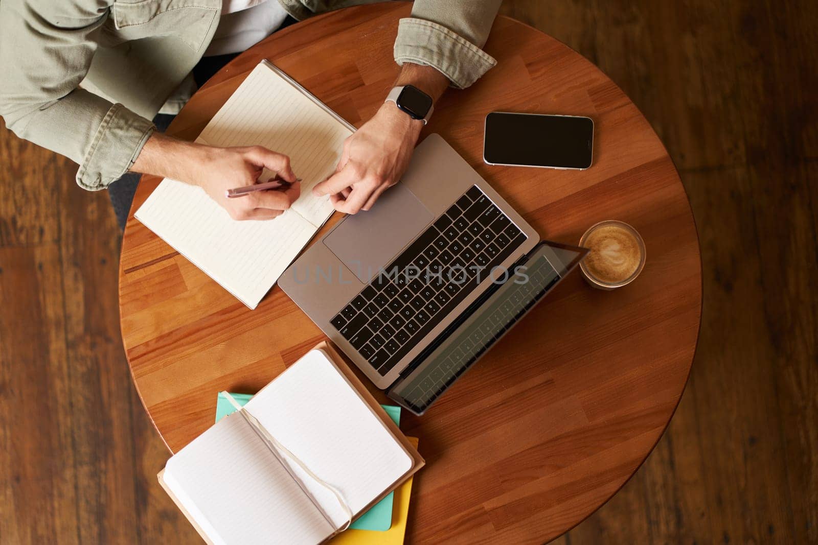 Top aerial view of male hands taking notes, writing in notebook, man sitting at round desk in cafe, working on laptop, studying.