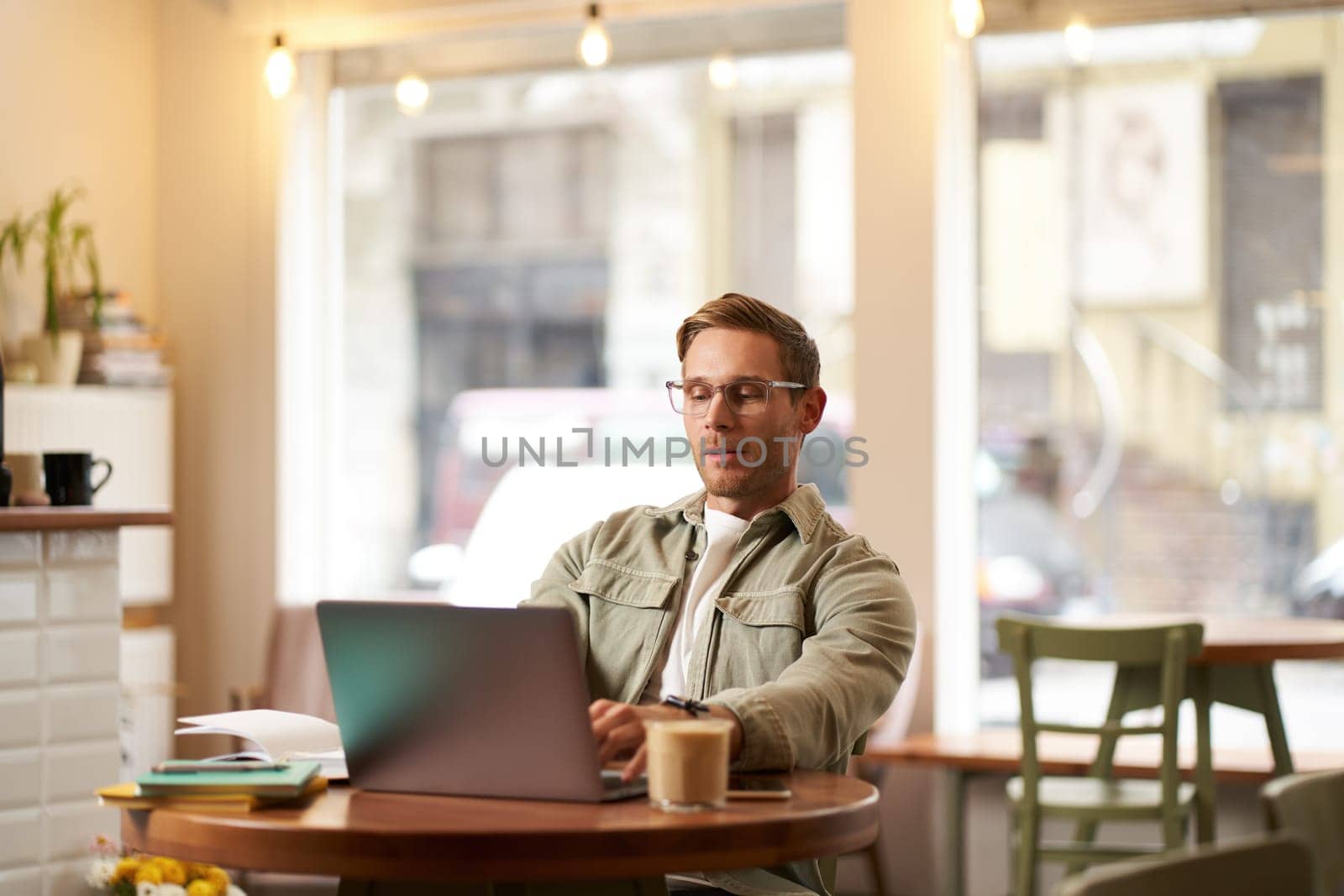 Portrait of man with serious face, sitting in cafe, wearing glasses, working on laptop, looking concentrated, doing freelance project.