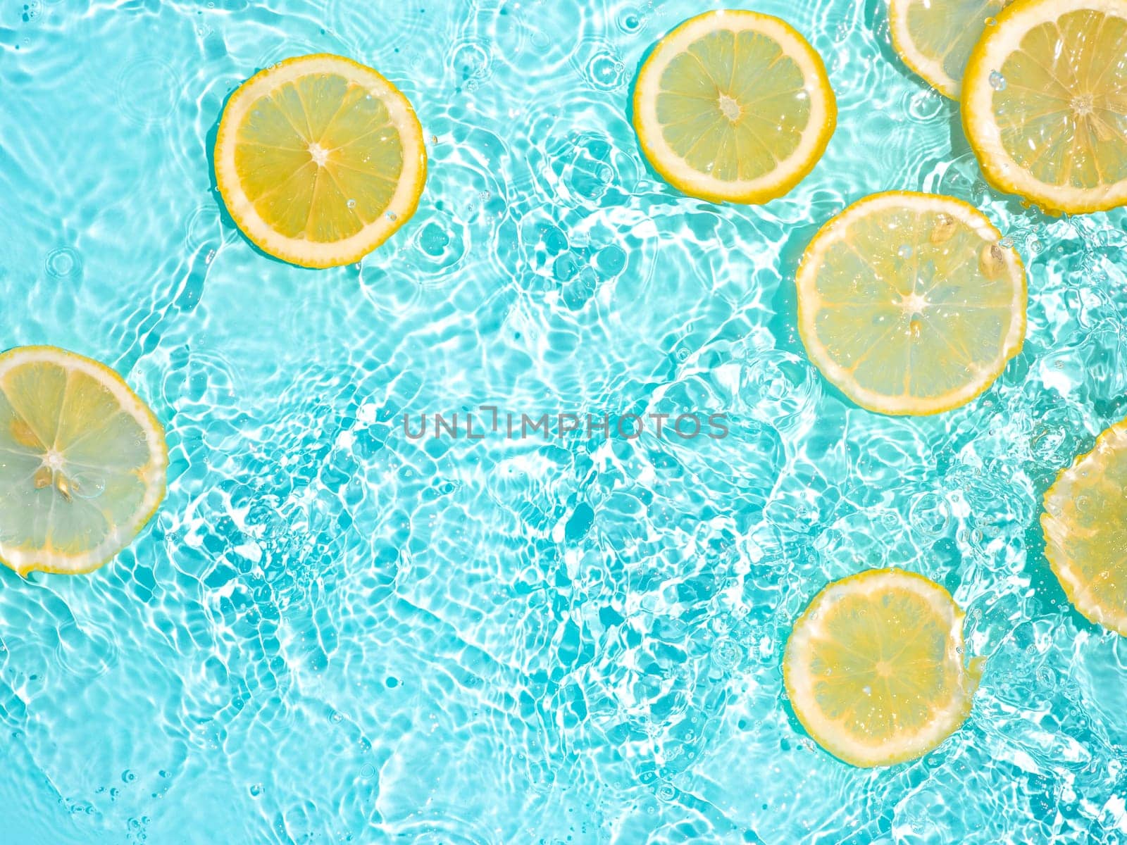 Lemon slices in clean transparent water over blue background with copy space. Water splashing on blue water surface in sunlight. Top view or flatlay. Summer, vacation, healthy eating concept
