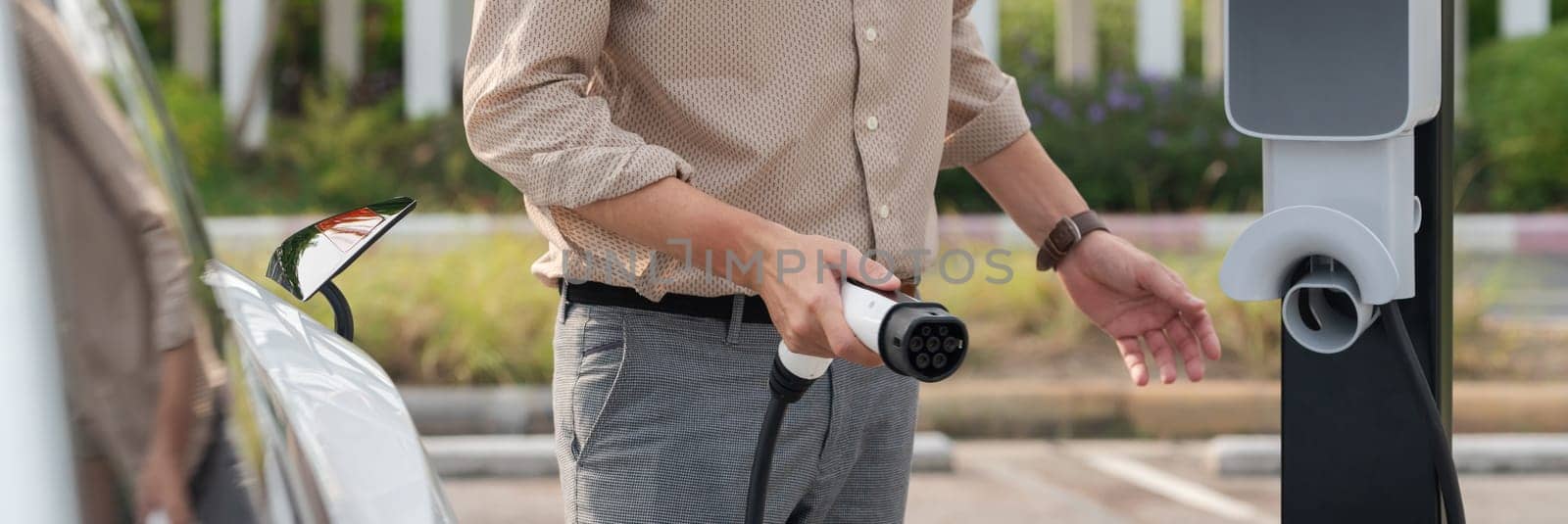 Young man recharge electric car's battery from charging station in city commercial parking lot. Rechargeable EV car for sustainable environmental friendly urban travel. Panorama Expedient