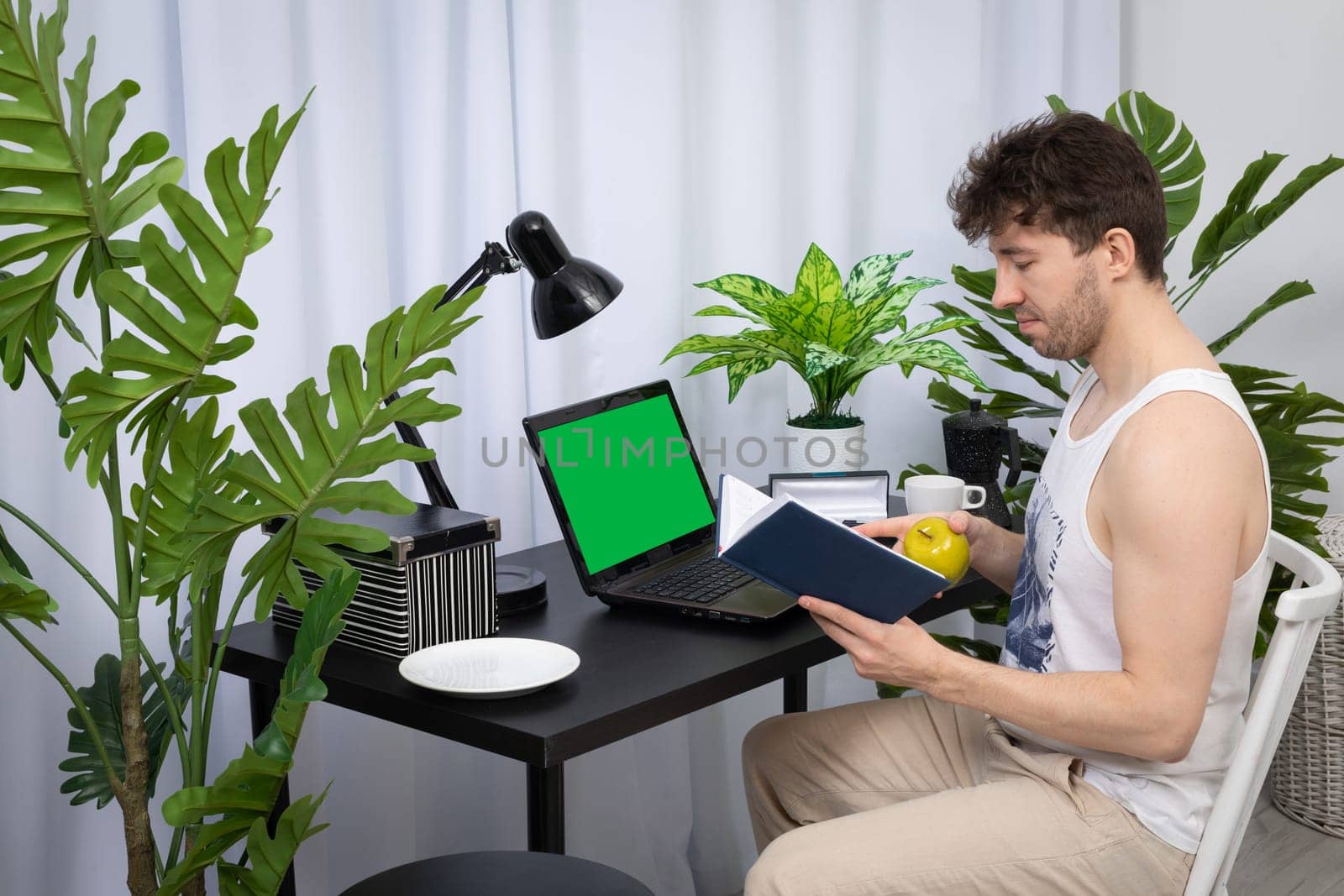Sitting at the desk at home, the employee checks the entries in the book calendar to know where to start work. Remote work during a pandemic. A bright living room in a modern style with a preferable space. Green screen of the monitor.
