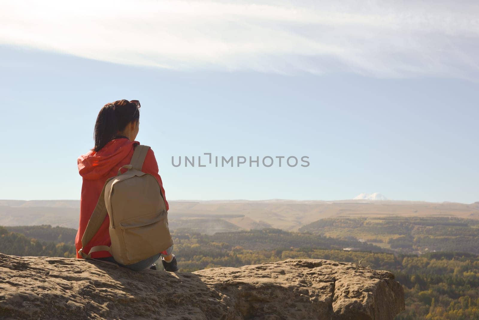 A girl tourist with a backpack sits on the top of a mountain and enjoys the beautiful views. A walking woman in an orange jacket rests on a rock, looking at the beautiful sunny landscape of the Caucasus Mountains