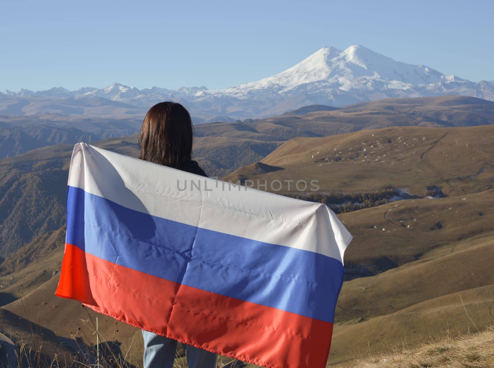 A young brunette woman stands against the backdrop of the snow-capped Mount Elbrus, looking at the beauty of the mountains, holding a Russian flag in her hands. Tricolor against the backdrop of snow-capped Mount Elbrus by Ekaterina34
