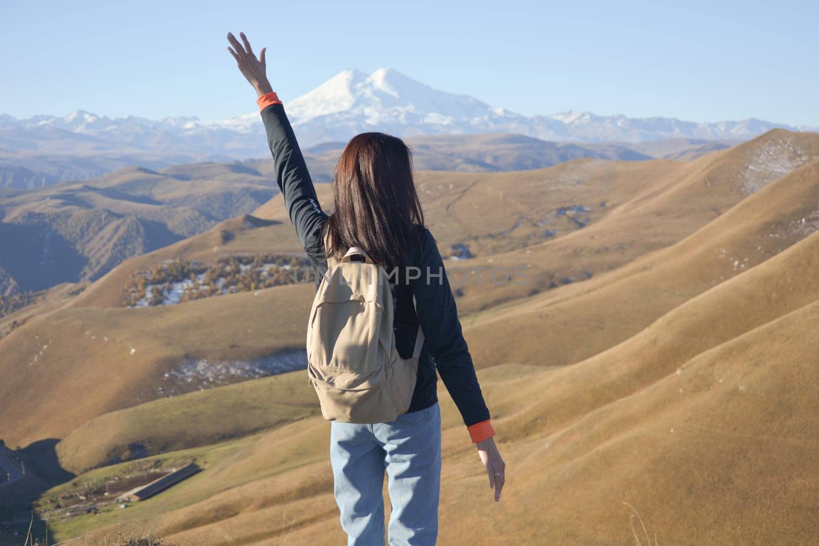 A young brunette woman with a backpack stands against the background of snow-capped Mount Elbrus and looks towards the mountains