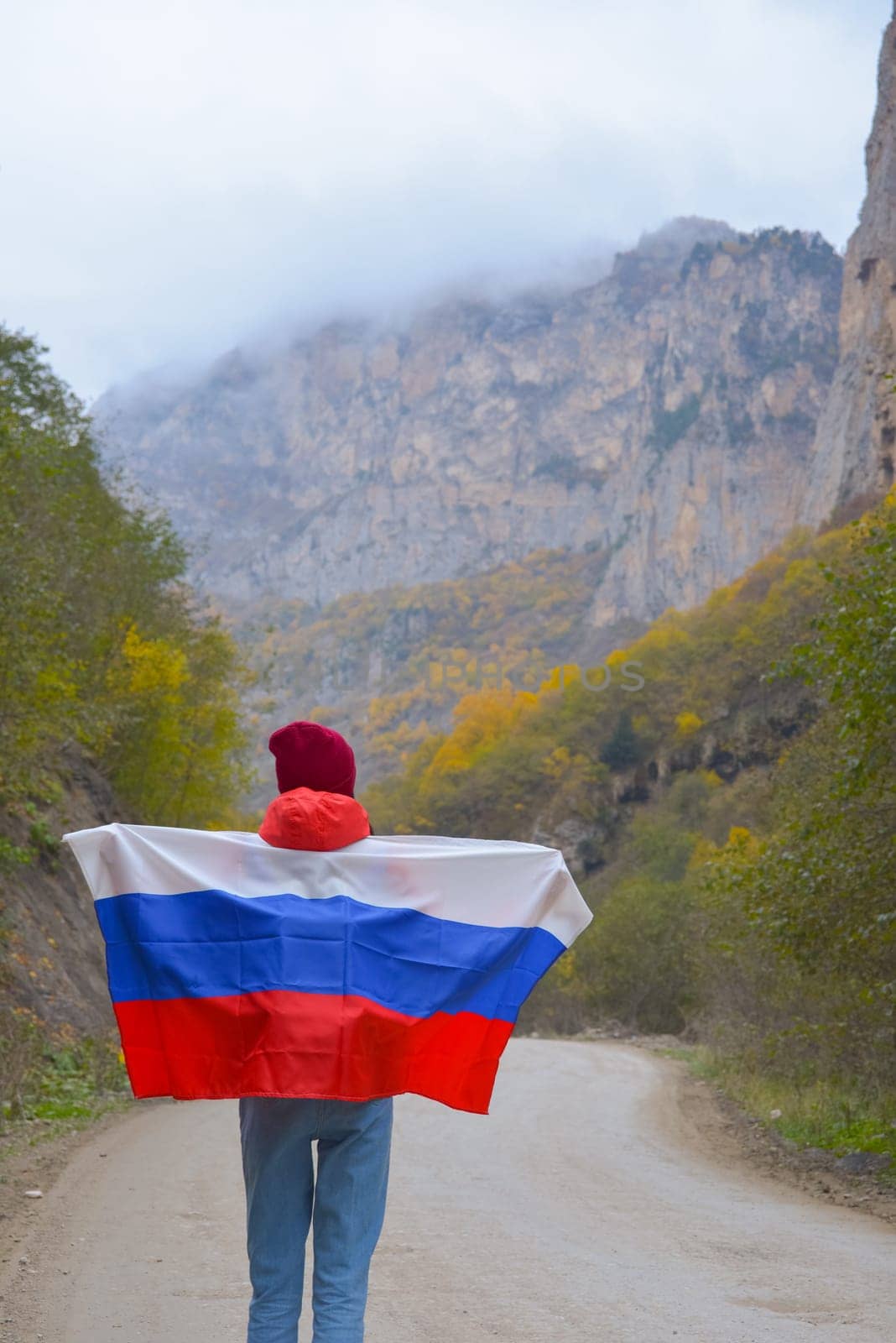 Woman traveler on a winding road in the mountains on a foggy autumn day with the flag of the Russian Federation on her shoulders.