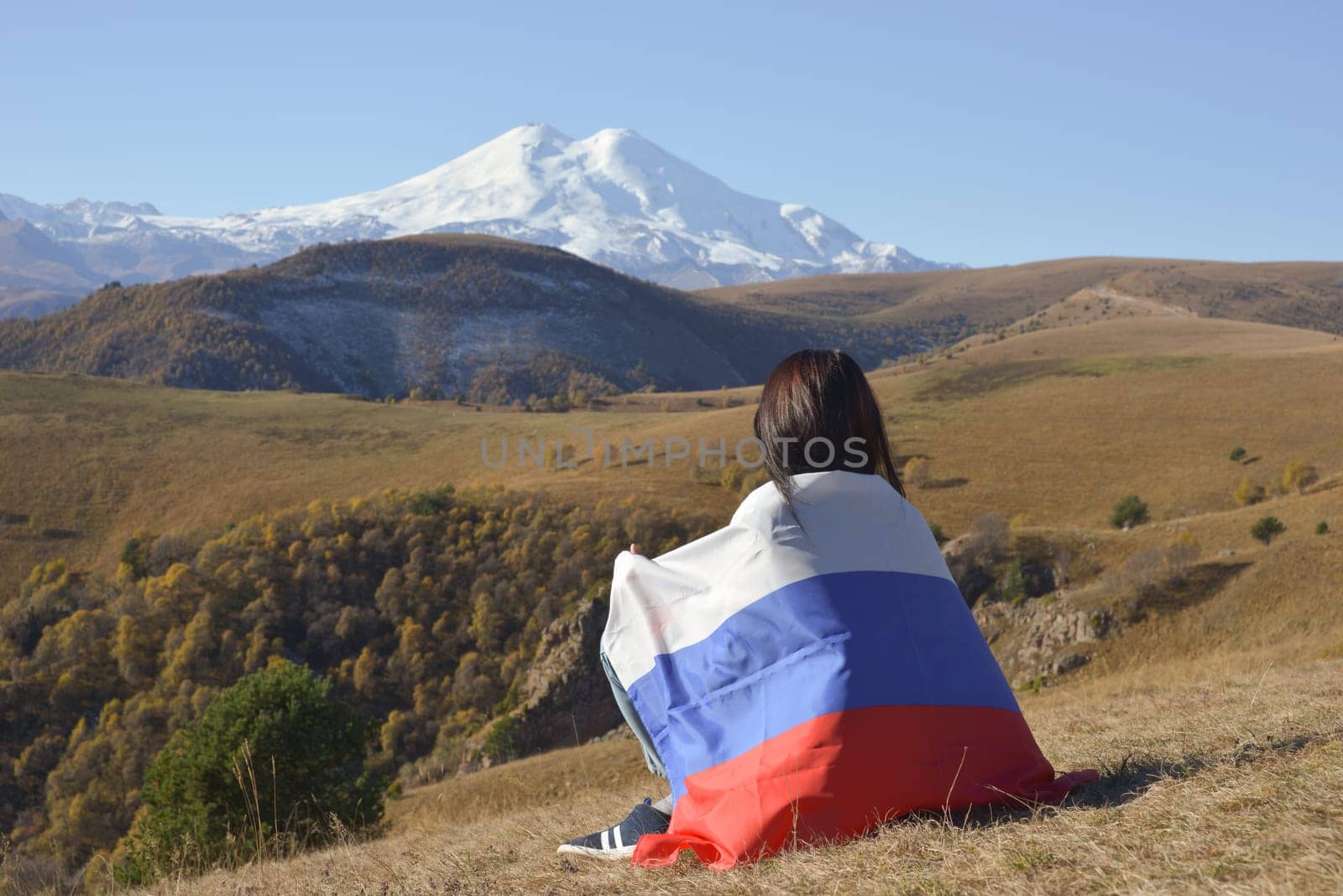A young brunette woman sits on a stone and looks at the mountains around, her shoulders are covered with a Russian flag. Against the backdrop of snow-capped Mount Elbrus by Ekaterina34