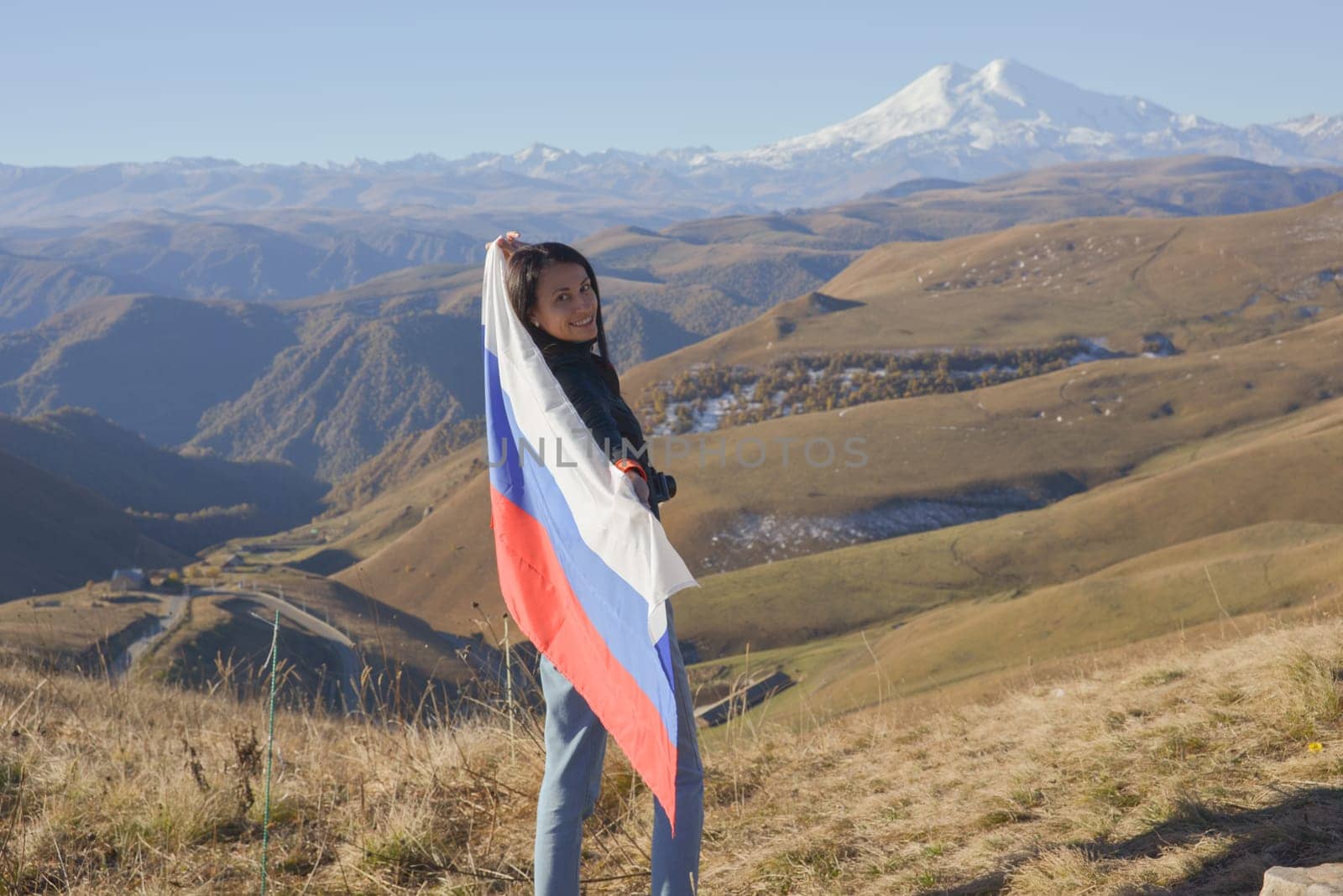 A young brunette woman stands against the backdrop of the snow-capped Mount Elbrus, looking at the camera, a Russian flag covers her shoulders. Tricolor against the backdrop of snow-capped Mount Elbrus.