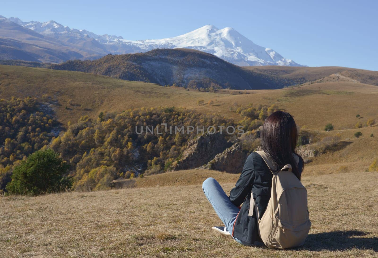 A female traveler with a backpack behind her sits on the grass and enjoys the views of Elbrus. View of Elbrus, North Caucasus, Russia