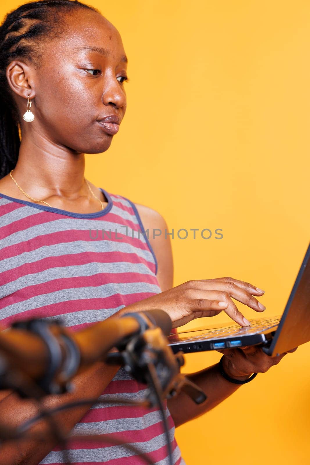 Dedicated young lady engrossed in inspecting and servicing bicycle with laptop. African American woman resolves broken bike with digital pc, ensuring safety and functionality. Close up shot.