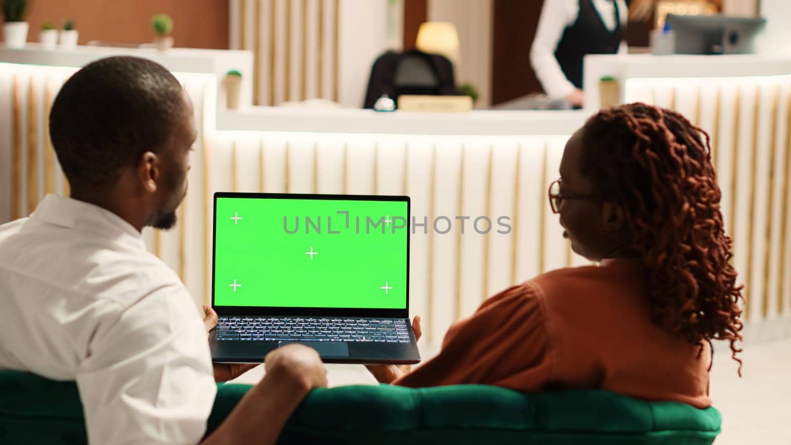 Happy couple on their honeymoon vacation holding laptop chroma key green screen mock up, talking with family in video call. Husband and wife chatting in video conference sitting in hotel lobby