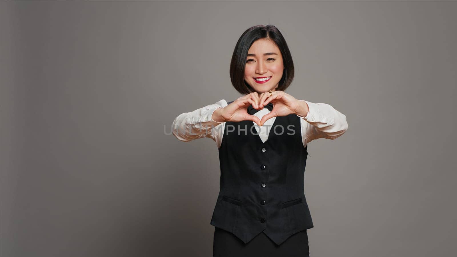 Hotel administrator shows heart shape symbol on camera, expressing romantic gesture and being flirty. Asian receptionist presenting true honest feelings, does romance sign in studio. Camera A.