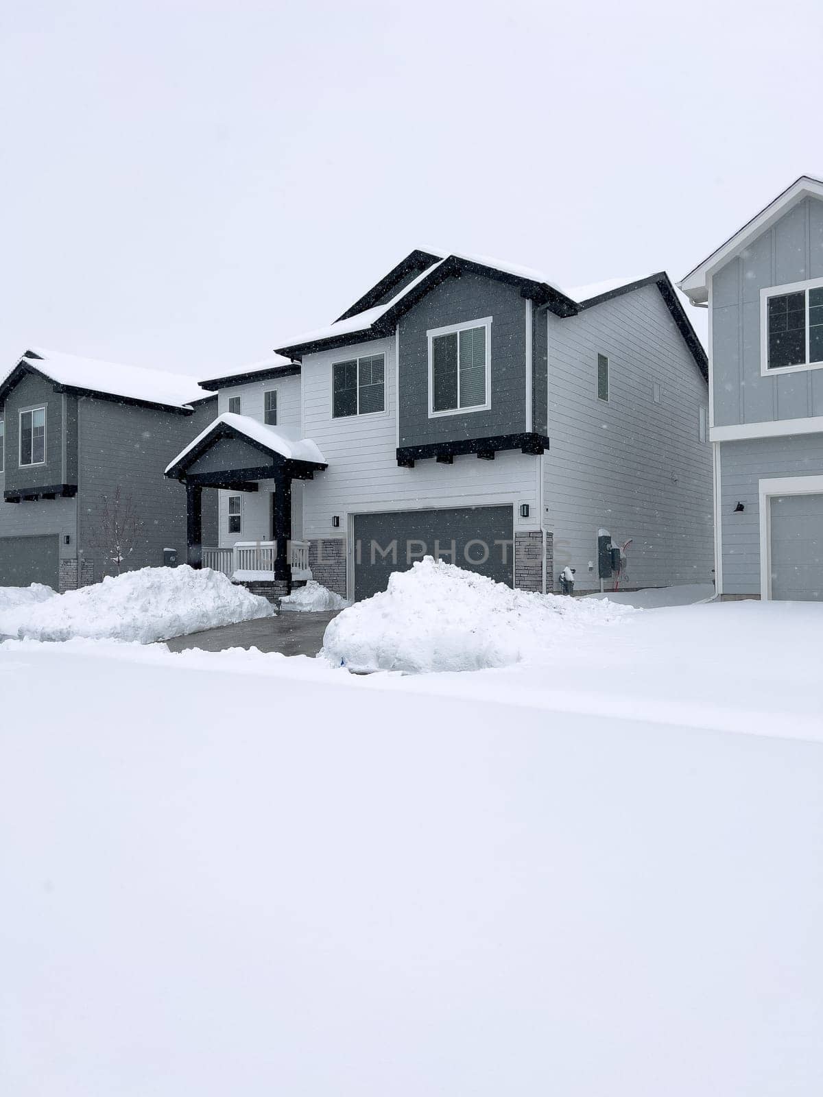 The scene depicts a suburban tranquility with homes lining a street blanketed in snow, punctuated by a cleared path that weaves through the winter wonderland. The overcast sky promises more snow, creating a hushed atmosphere over the neighborhood.