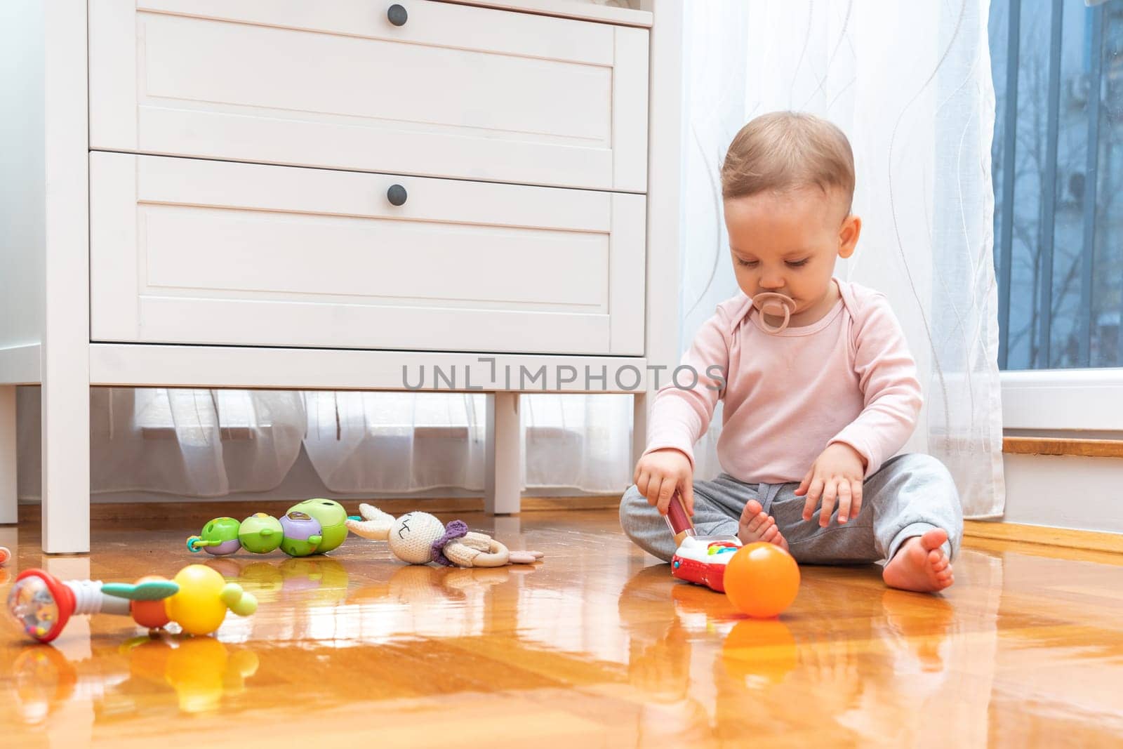 A baby with a pacifier in his mouth interacts with interest with toys while playing in the room.