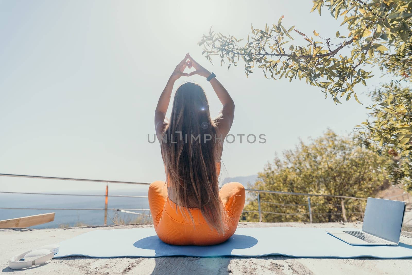 Fitness woman sea. Happy middle aged woman in orange sportswear exercises morning outdoors on yoga mat with laptop in park over ocean beach. Female fitness pilates yoga routine. Healthy lifestyle. by panophotograph