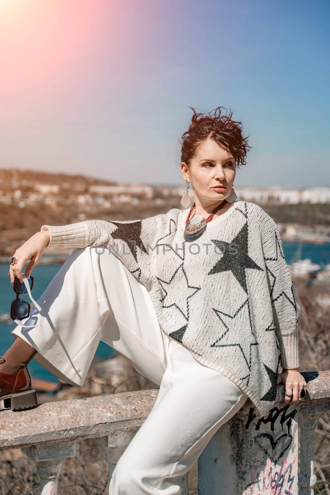 Woman walks around the city, lifestyle. A young beautiful woman in white trousers and a sweater sits on a white fence with balusters and overlooks the sea bay and the city. by Matiunina