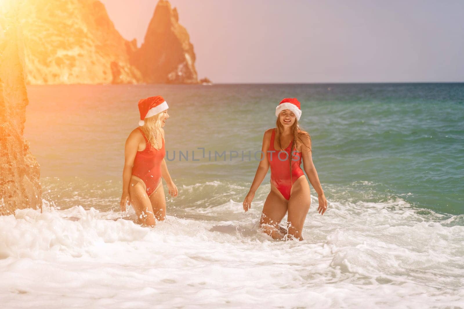 Women Santa hats ocean play. Seaside, beach daytime, enjoying beach fun. Two women in red swimsuits and Santa hats are enjoying themselves in the ocean waves