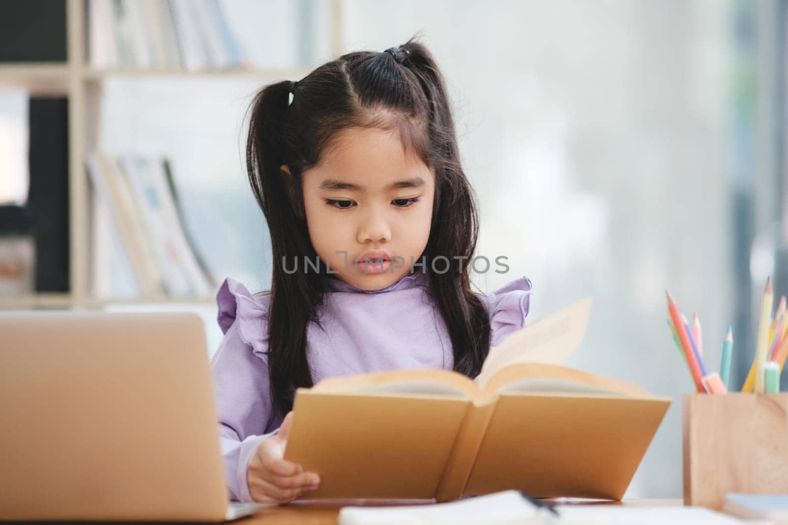 A young girl is sitting at a desk reading a book. She is wearing a purple shirt and has her hair in pigtails. The scene is set in a library or a classroom, with a laptop and a pencil on the desk
