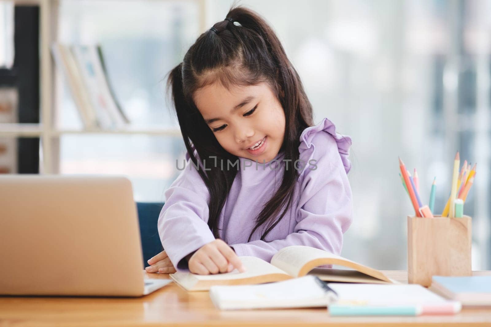 A young girl is sitting at a desk with a laptop and a book. She is smiling and she is enjoying her work
