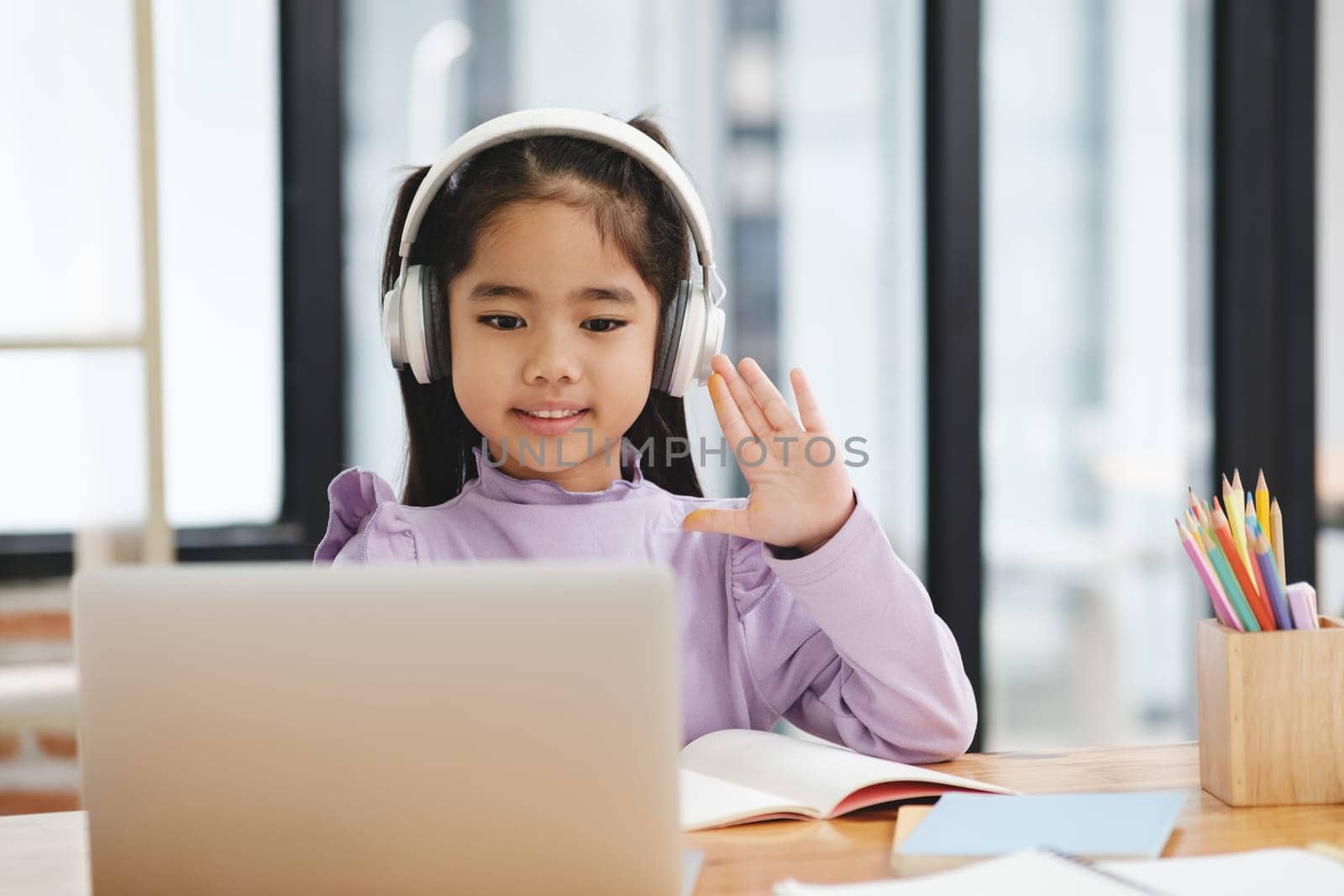 A young girl wearing headphones is waving at the camera while sitting at a desk with a laptop and a book