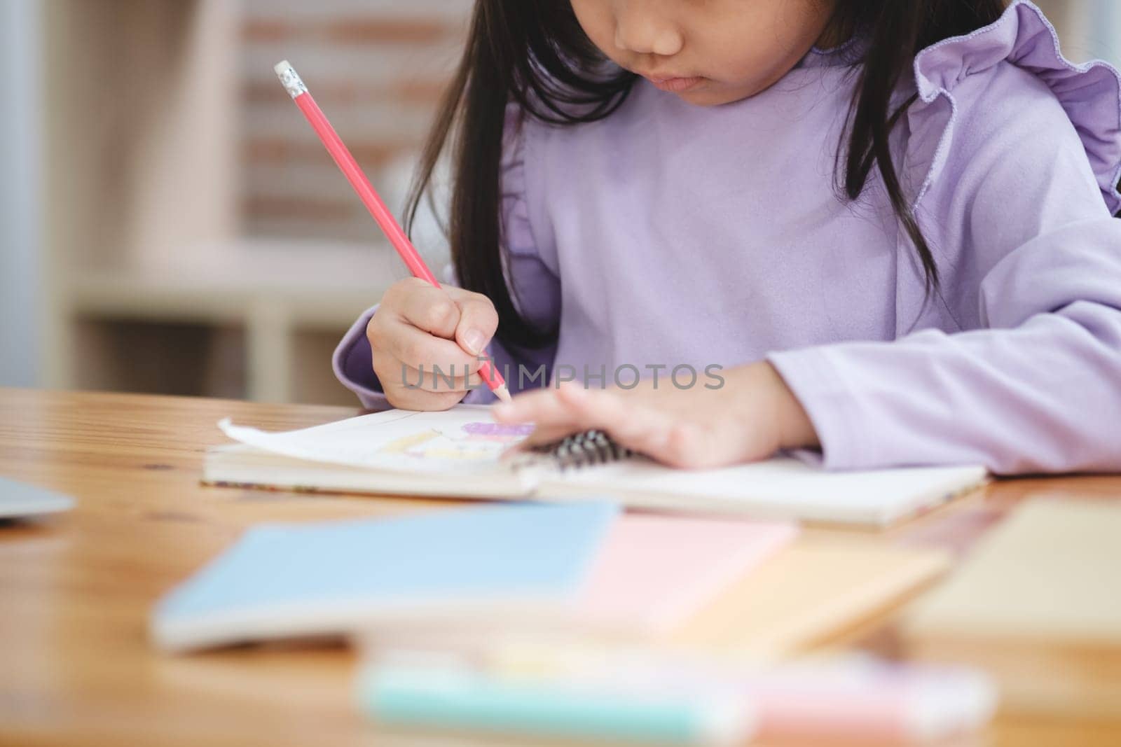 A young girl is writing with a red pencil on a piece of paper. She is sitting at a desk with a laptop and a stack of books nearby