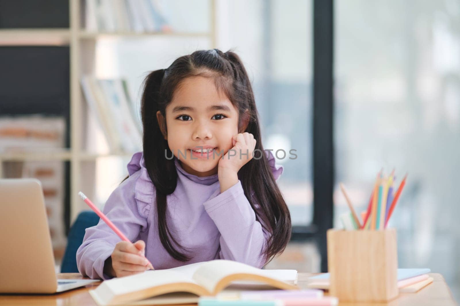 A young girl is sitting at a desk with a laptop and a book. She is smiling and she is enjoying herself