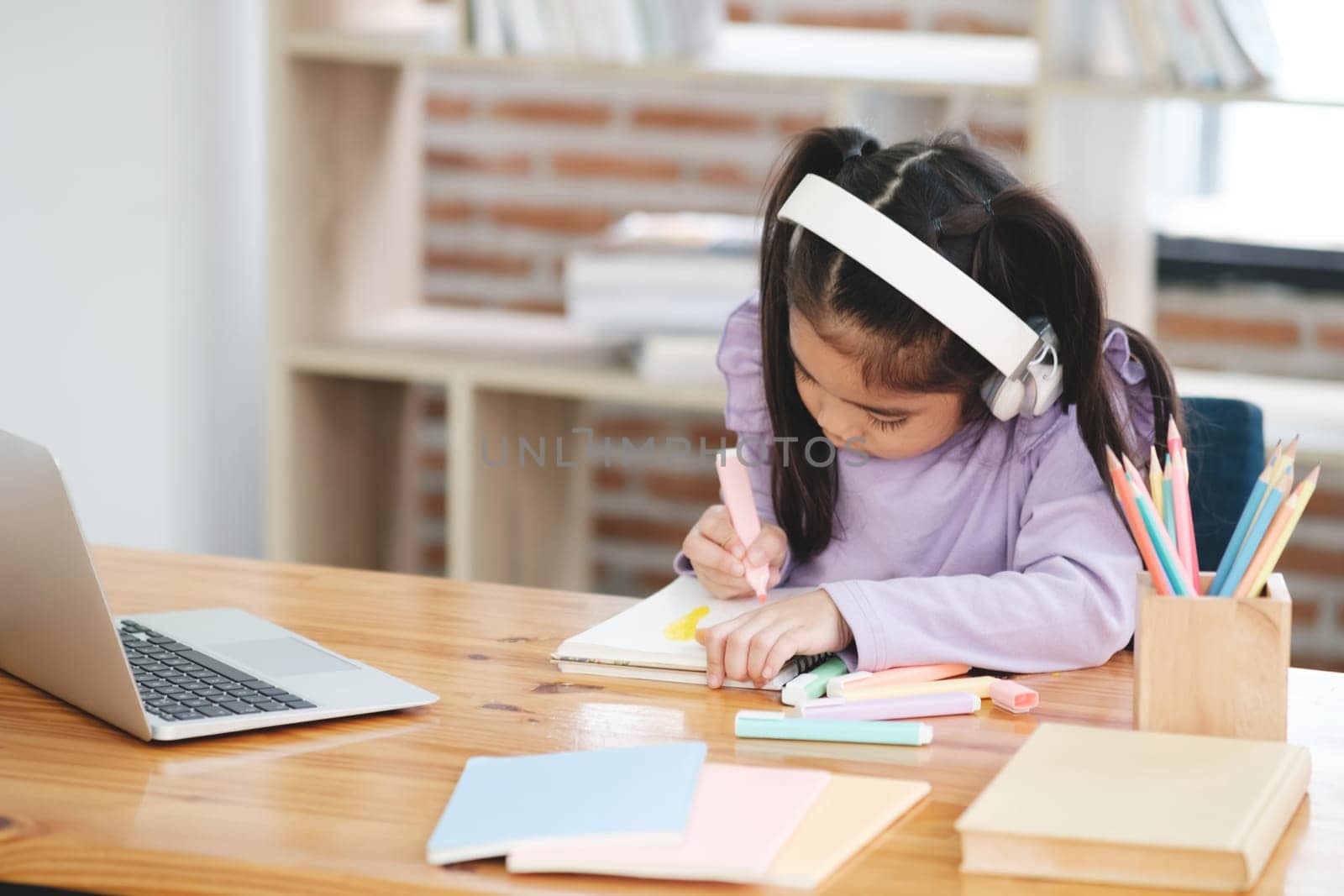 A young girl is sitting at a desk with a laptop and a notebook. She is drawing with markers and wearing headphones. The scene suggests that she is working on a project or assignment