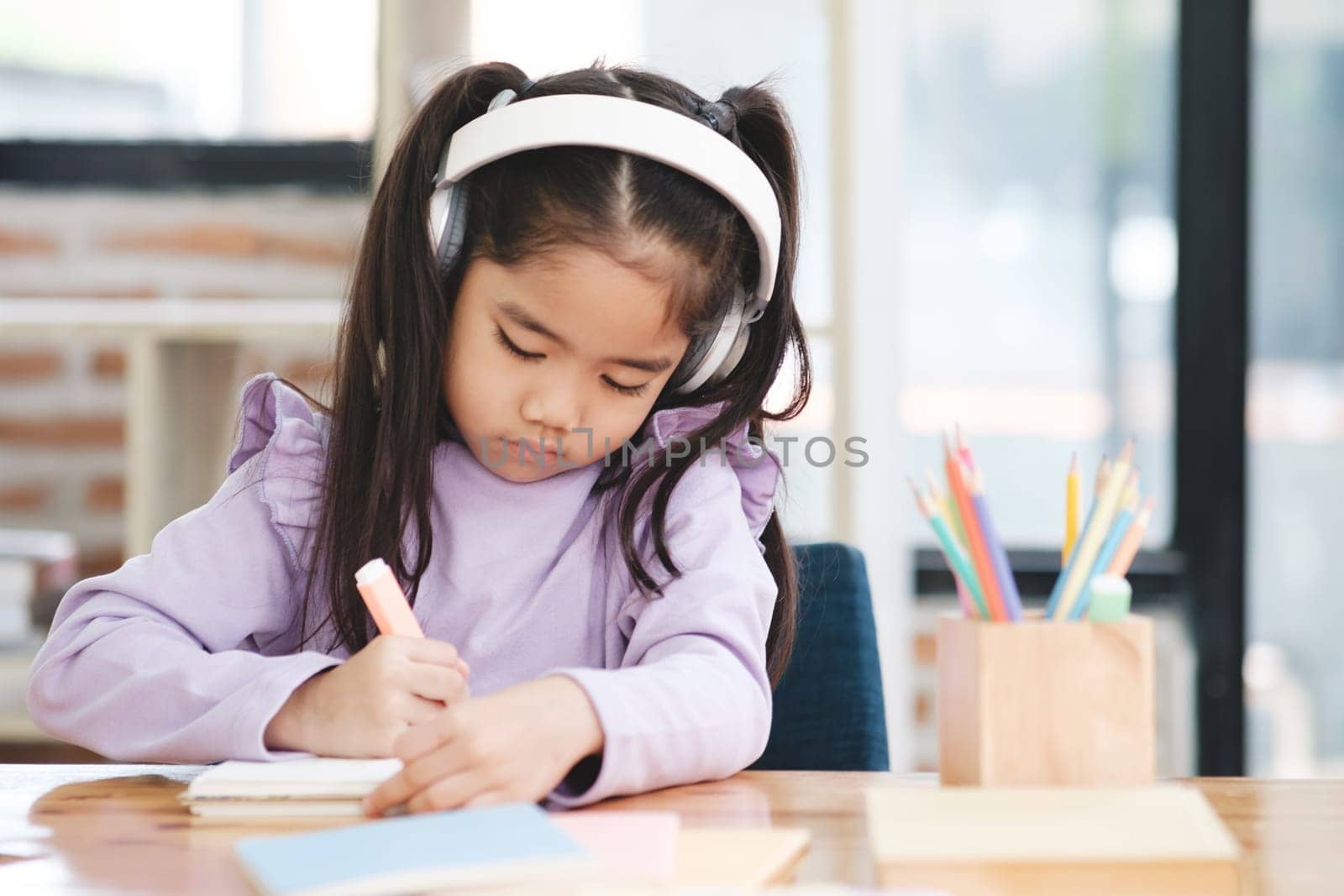 A young girl is sitting at a desk and writing in a notebook. She is wearing headphones and she is focused on her work. The scene suggests that she is engaged in a creative or educational activity