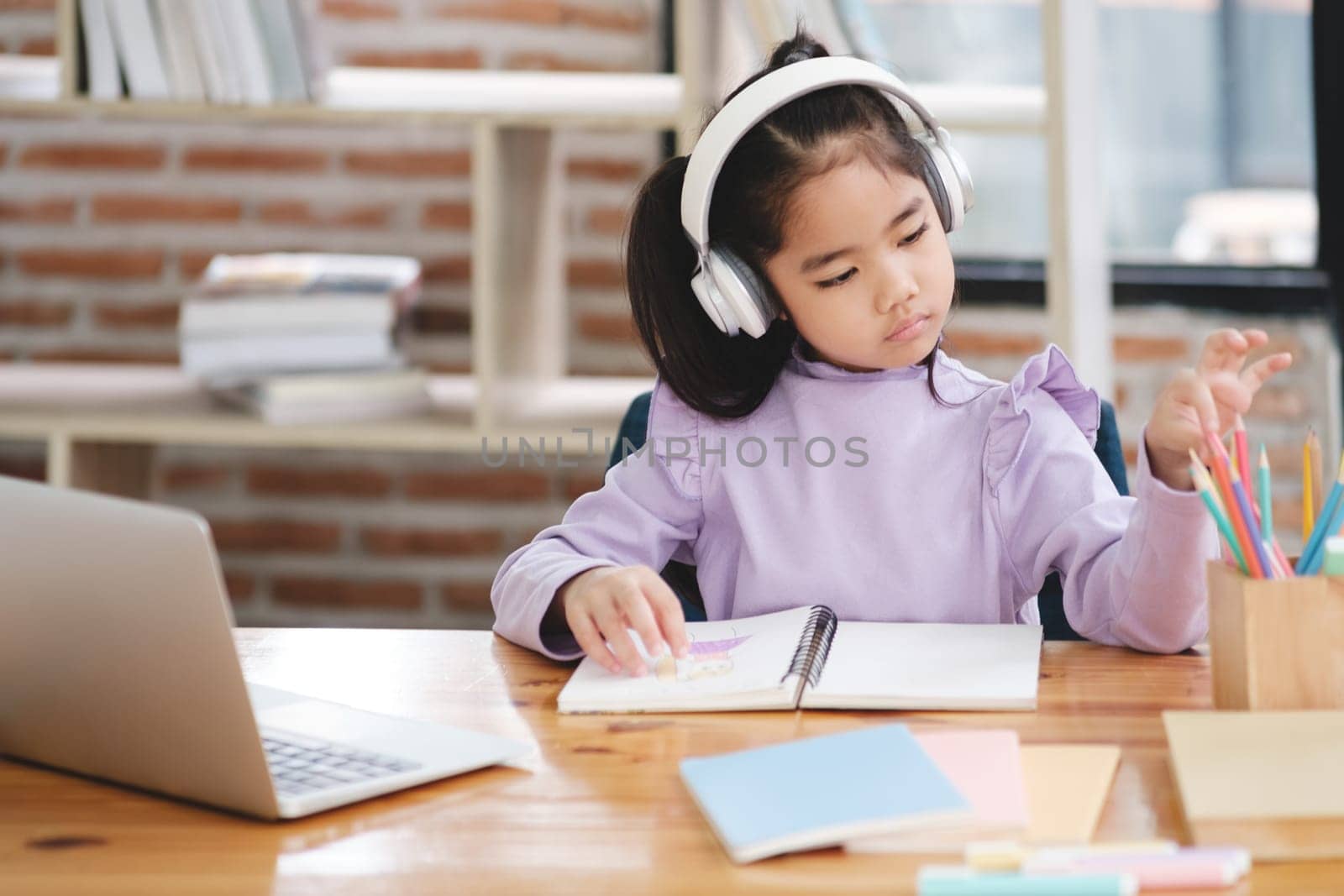 A young girl is sitting at a desk with a laptop and a notebook. She is wearing headphones and she is focused on her work