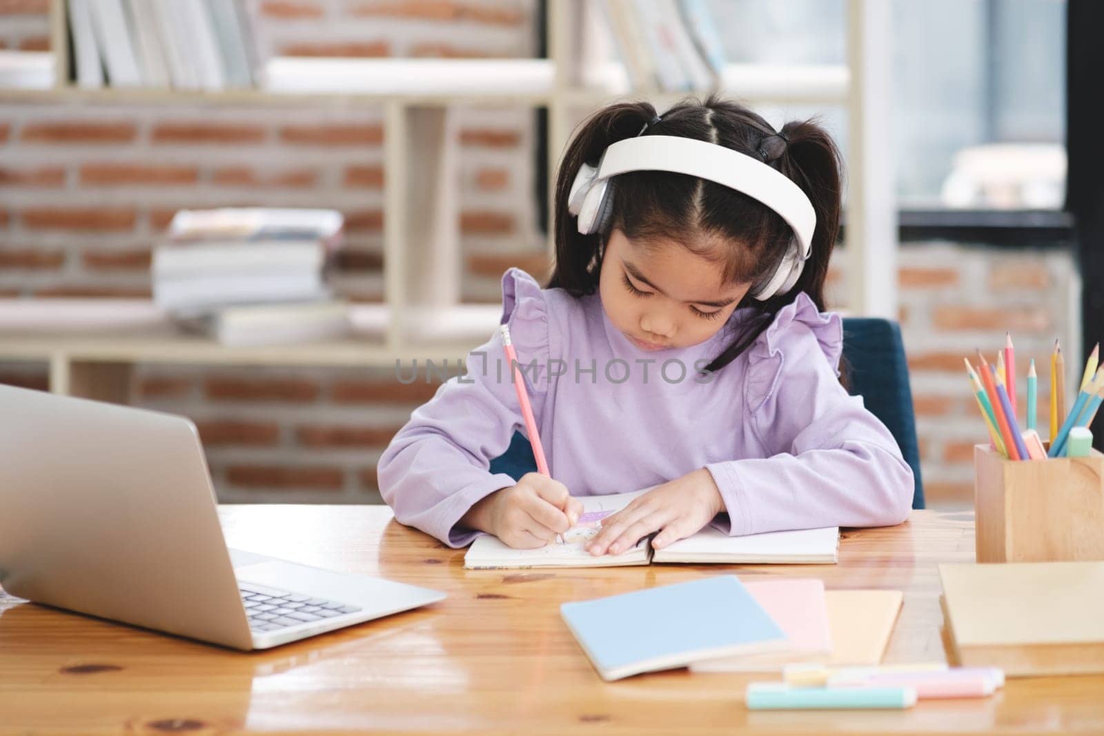 A young girl is sitting at a desk with a laptop and a notebook. She is writing in her notebook and wearing headphones. The scene suggests that she is working on a project or studying for an exam