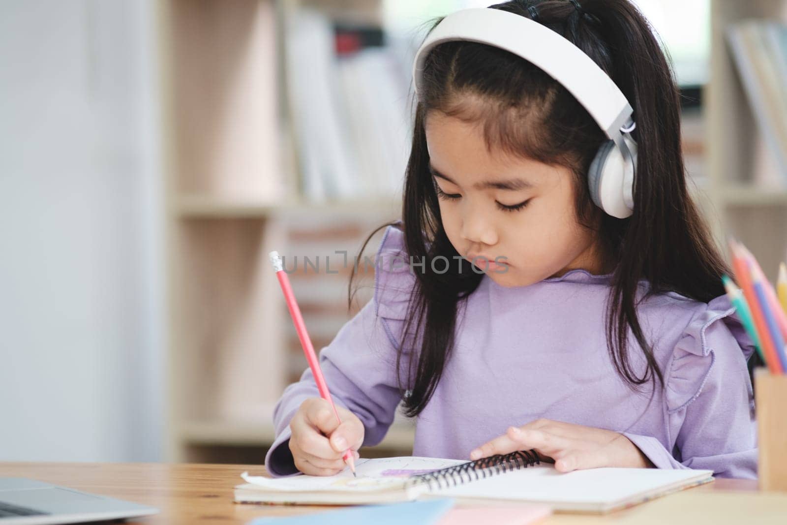 A young girl is sitting at a desk with a pencil and a notebook. She is wearing headphones and she is focused on her work. The scene suggests that she is engaged in a task that requires concentration