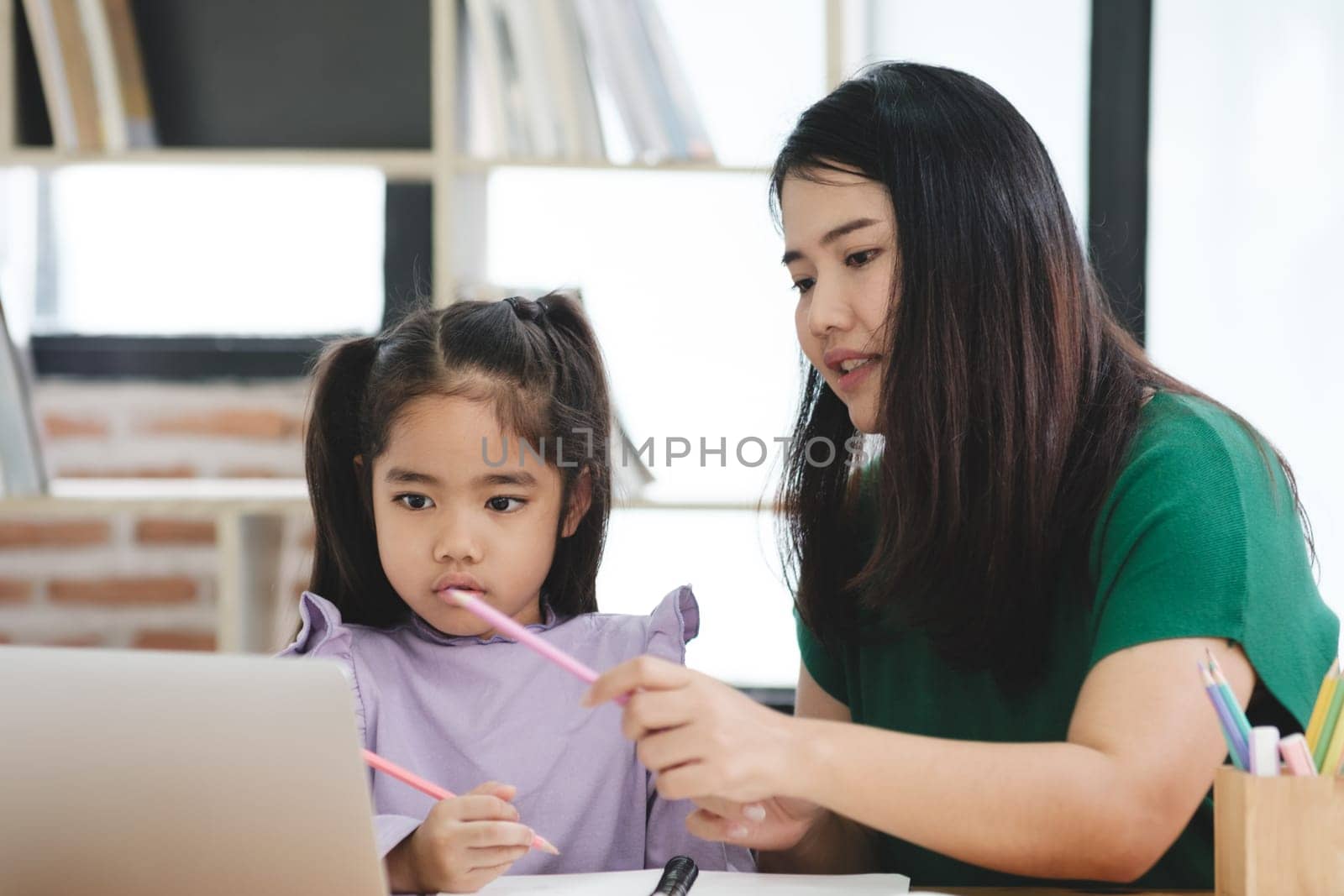 A woman is helping a young girl with her homework. The girl is using a pencil and the woman is holding a pen. The scene is set in a room with a lot of books and a laptop