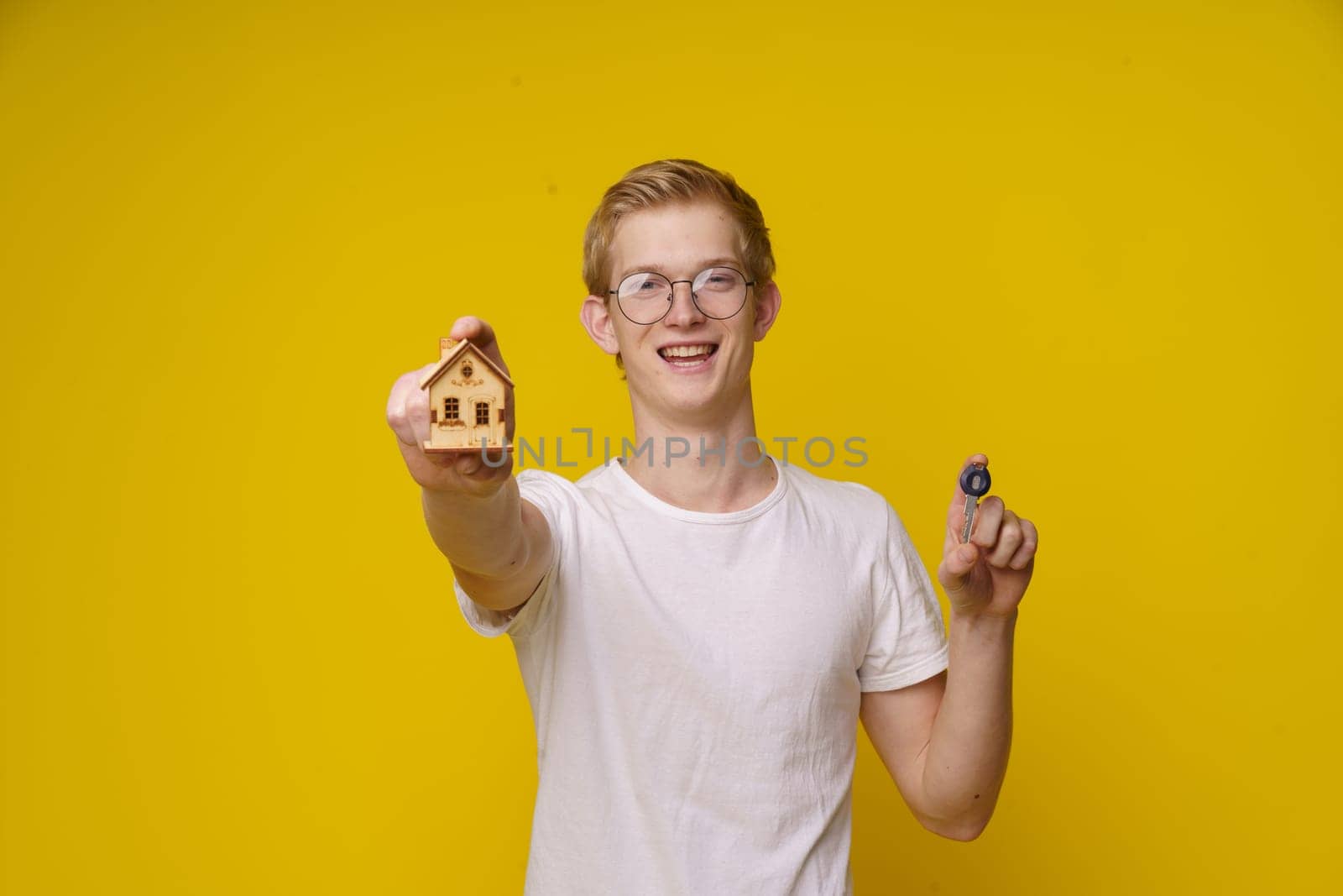 nerdy individual with glasses is holding a mock-up of a house and a key to housing on a bright yellow background. The focus is on the hand holding the house, representing the concept of real estate investment, homeownership, and financial success through hard work and education by LipikStockMedia