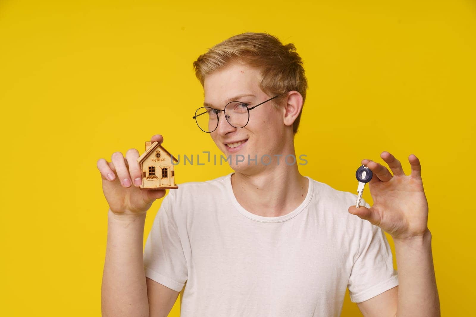 Young man with smile holds house model and home key in his hand, against yellow background with plenty of copy space. Concept of real estate, homeownership, rental, investment, or housing market. by LipikStockMedia