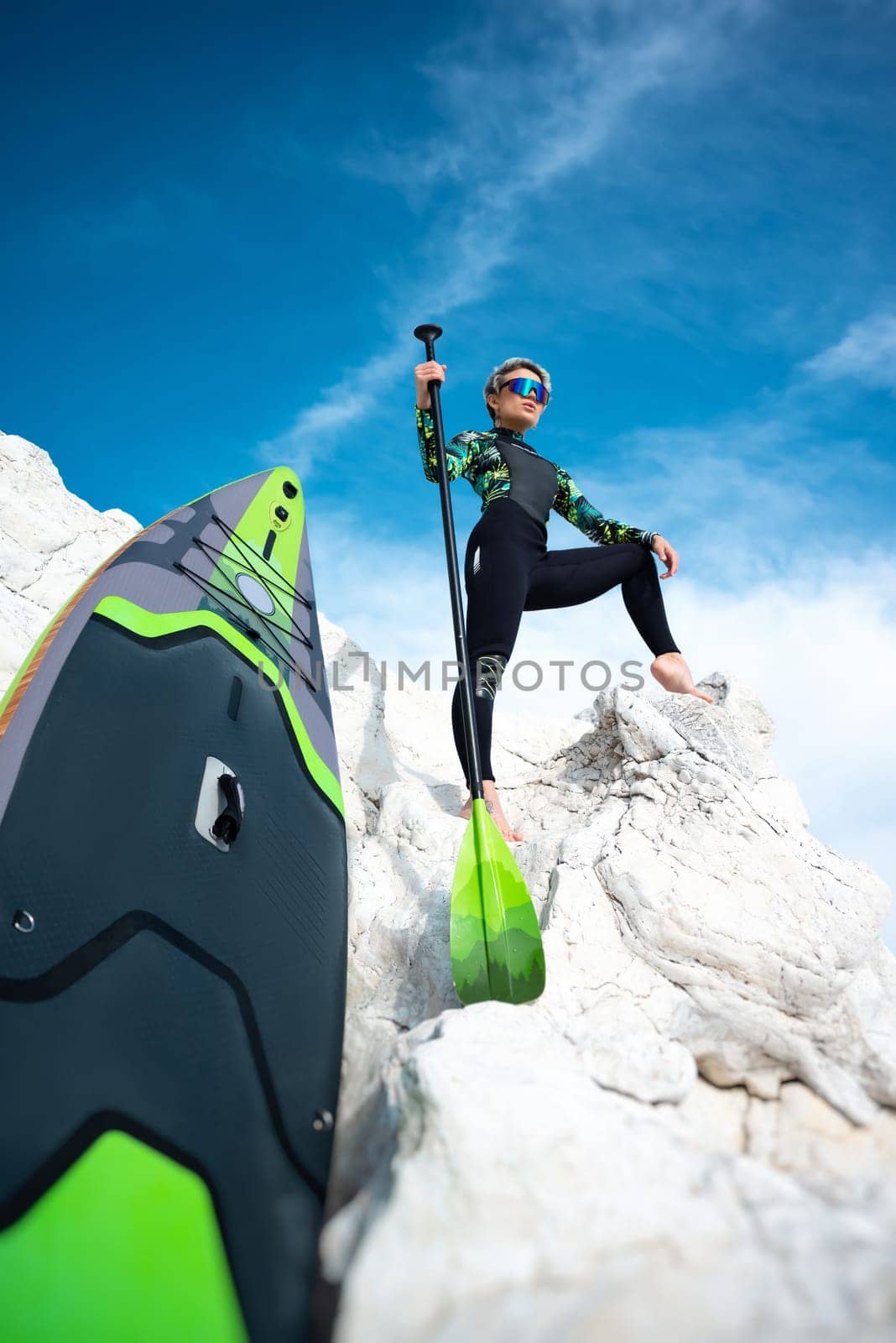 beautiful sup surfer girl in a wetsuit on the ocean shore against a beautiful blue sky poses sexually with a paddle