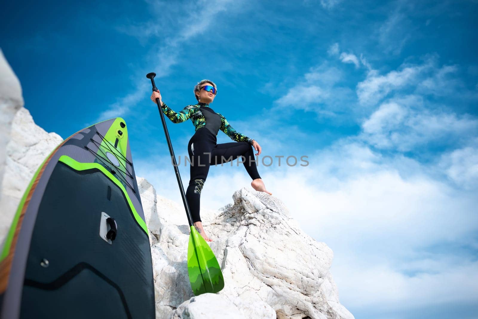 beautiful sup surfer girl in a wetsuit on the ocean shore against a beautiful blue sky poses sexually with a paddle