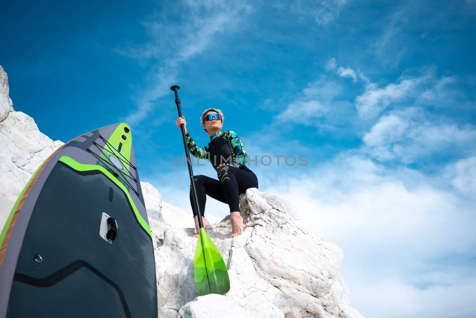 beautiful sup surfer girl in a wetsuit on the ocean shore against a beautiful blue sky poses sexually with a paddle