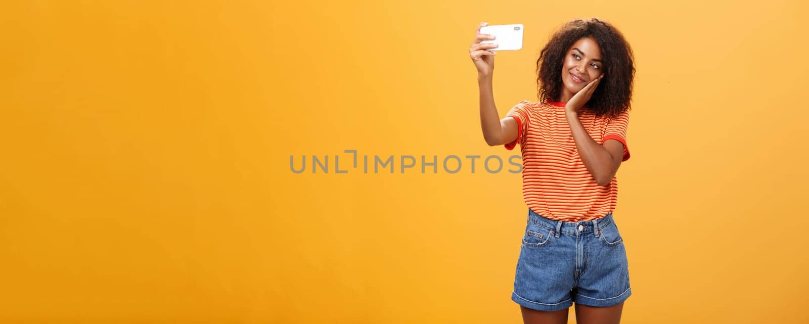 Woman made make-up all day to post new photo online taking selfie on brand new smartphone making cute face leaning head on palm smiling gently at device screen posing over orange background by Benzoix
