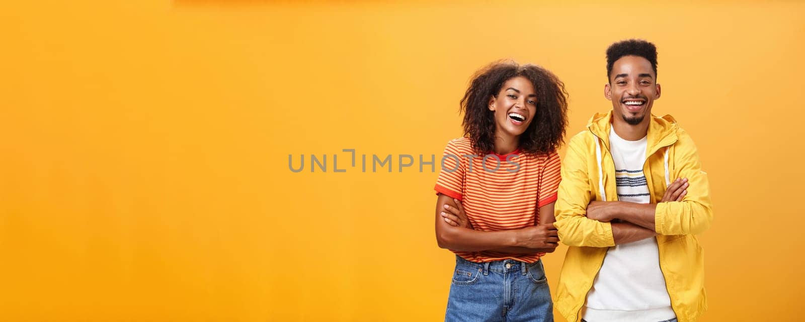 Two african american man and woman being best friends laughing out loud watching funny movie in cinema all dressed up in stylish outfit. standing with hands crossed on chest and amused expression.