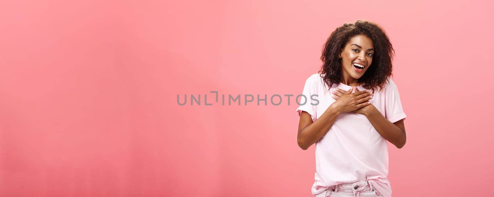 Portrait of charming delighted african american woman with curly haircut holding palms on heart pleased and grateful thanking friend for help smiling happily and thankful at camera over pink wall. Lifestyle.
